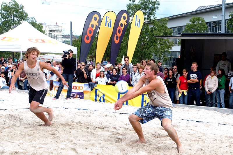 Zwei Männer spielen Beachvolleyball im Sand, umgeben von Zuschauern und Beachflags von Coop und Fa.