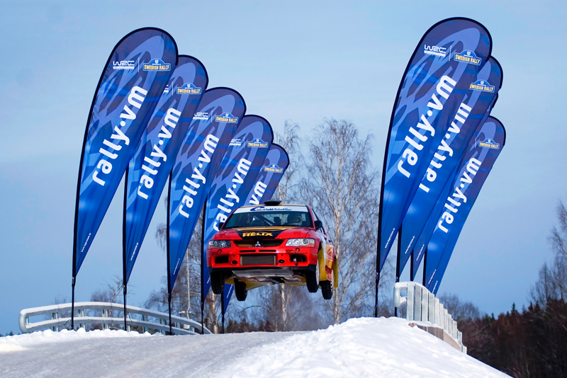 Ein Rallye-Auto springt über eine Schnee- und Eisfläche, umgeben von Beachflags mit dem Schriftzug 'rally.vum'.