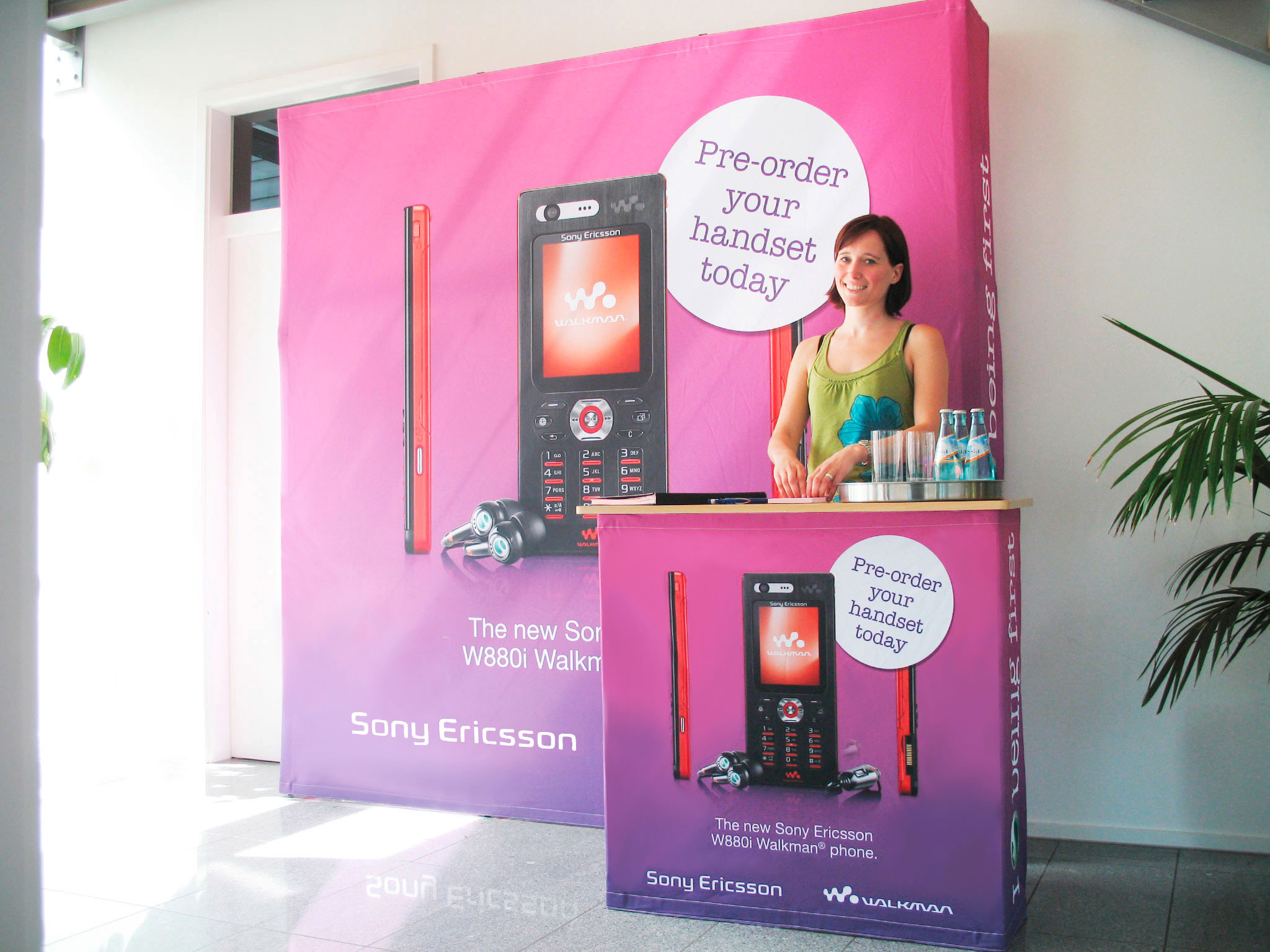 Messewand mit dem Produktnamen 'Quickup Stoffwand',  zeigt eine Frau hinter einer Messetheke mit Werbung für ein Sony Ericsson Handy