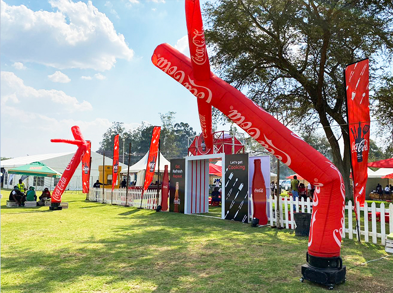Aufblasbare Werbung in Form von roten, tanzenden Figuren mit Coca-Cola Branding auf einem Eventgelände.