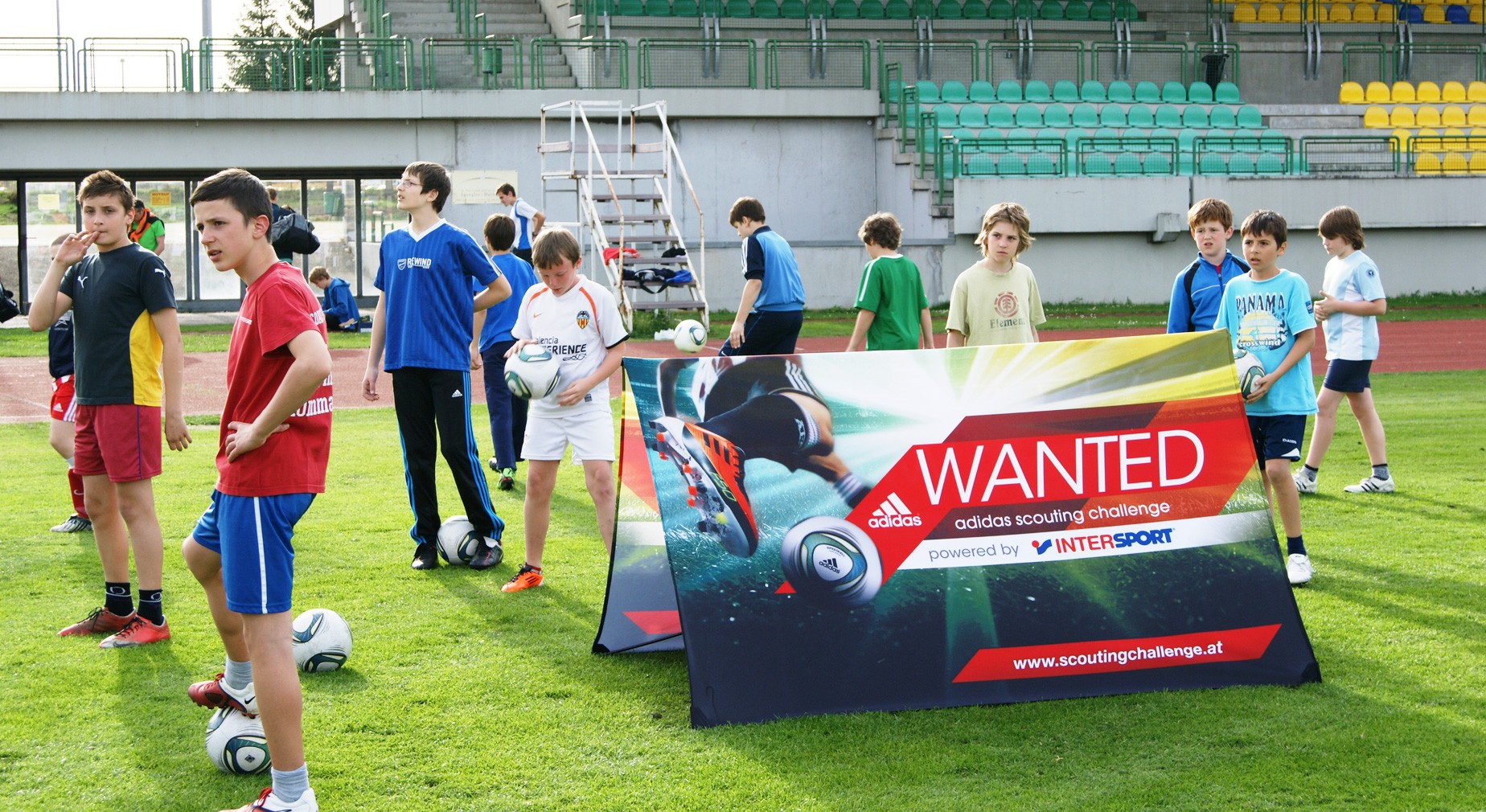 Gruppe von Kindern auf einem Sportplatz mit einer Werbebande für den 'adidas scouting challenge', die von Intersport unterstützt wird.