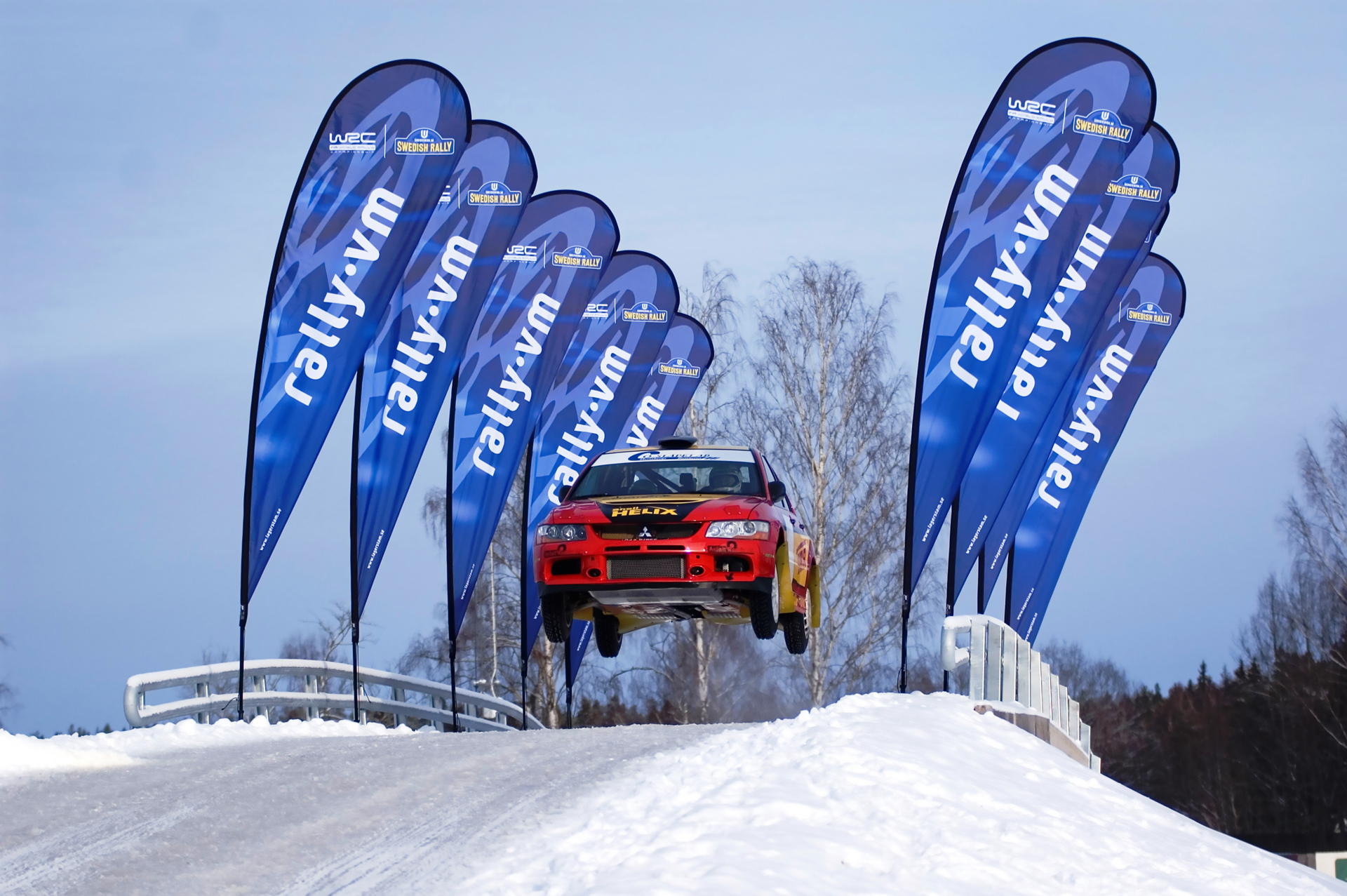 Ein Rallye-Auto springt über eine Schneehügel, umgeben von blauen Fahnen mit dem Schriftzug 'rally.vm'.