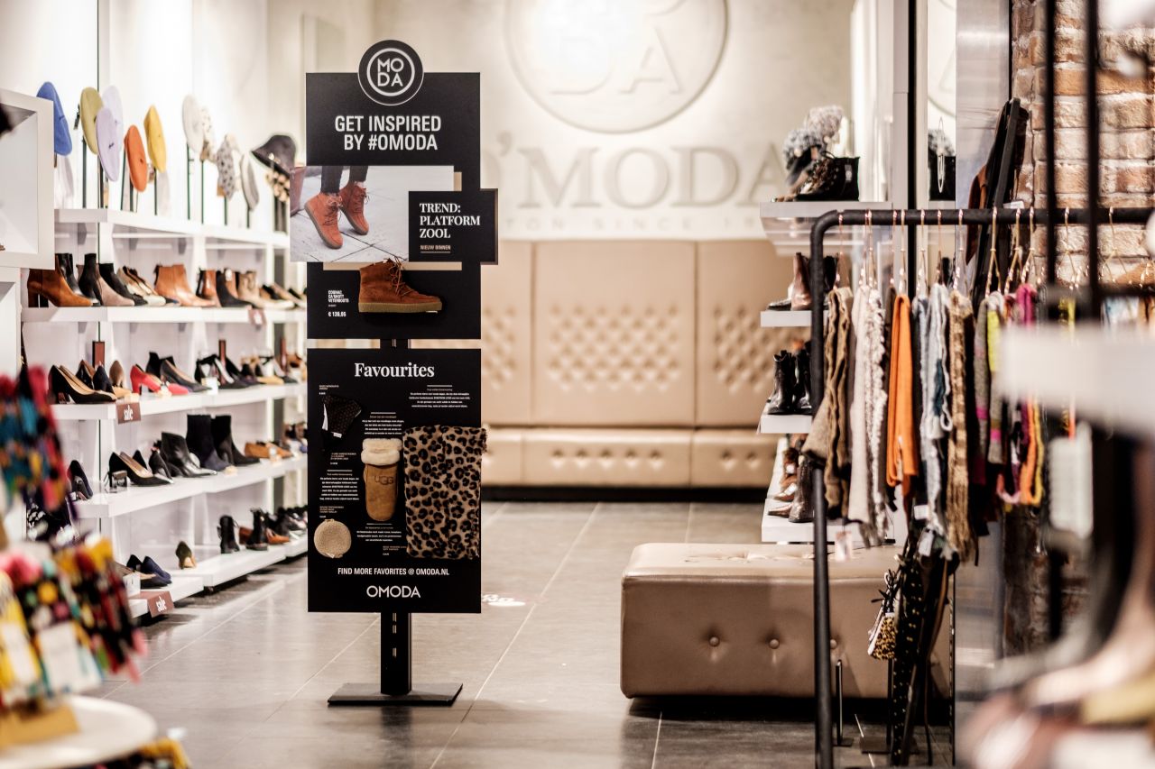 Inhaker POS Display in einer Modeboutique mit Schuhen und Kleidung im Hintergrund.