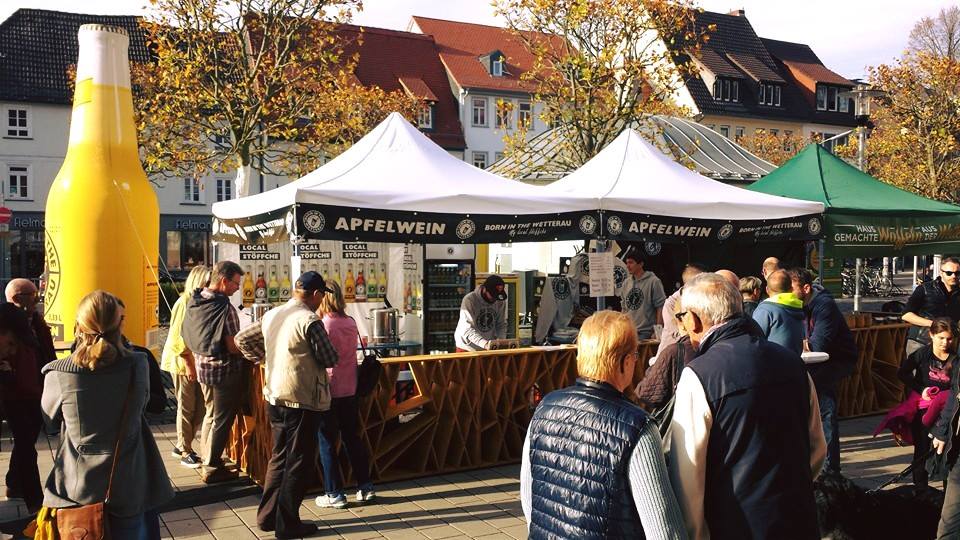 Marktstand mit aufblasbarer Flasche und Menschenmenge, die Apfelwein probiert.