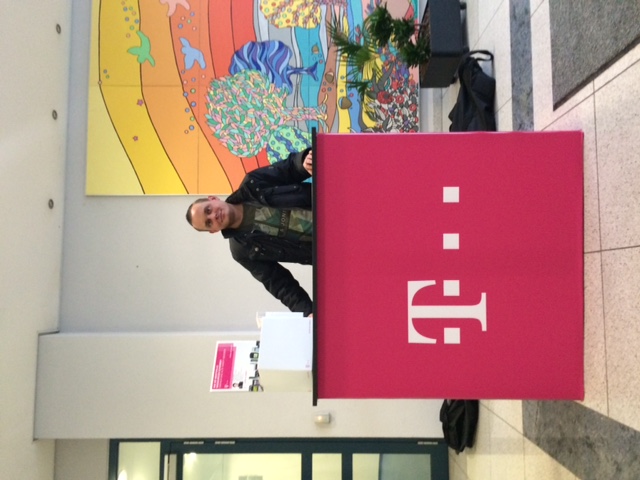 Mann steht an einem pinken Empfangstresen mit dem Logo von Telekom im Hintergrund. Bunte Wandmalerei sichtbar.