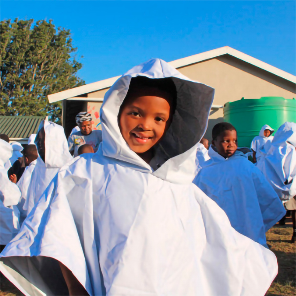 Ein lächelndes Kind in einem weißen Poncho steht im Vordergrund, umgeben von weiteren Kindern in ähnlicher Kleidung, vor einem Gebäude und einem Wassertank.