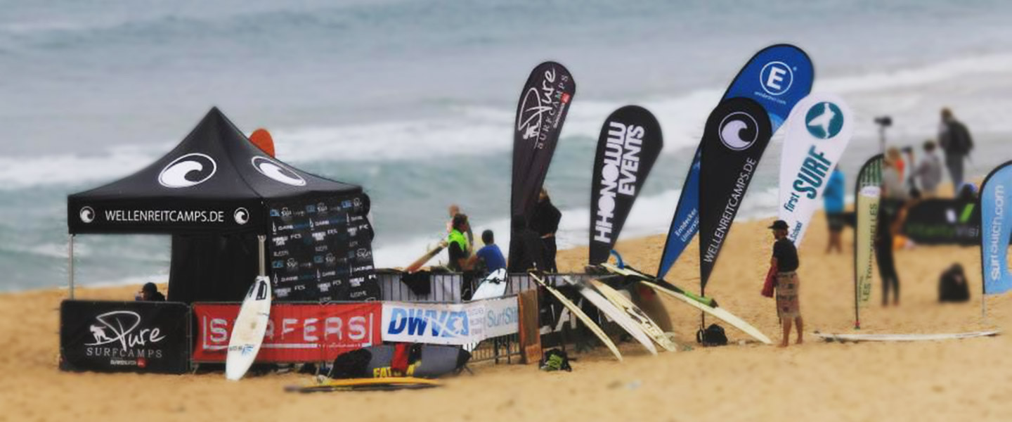 Zelt und Beachflags am Strand für das Wellenreitcamp, mit Surfboards und Teilnehmern im Hintergrund.
