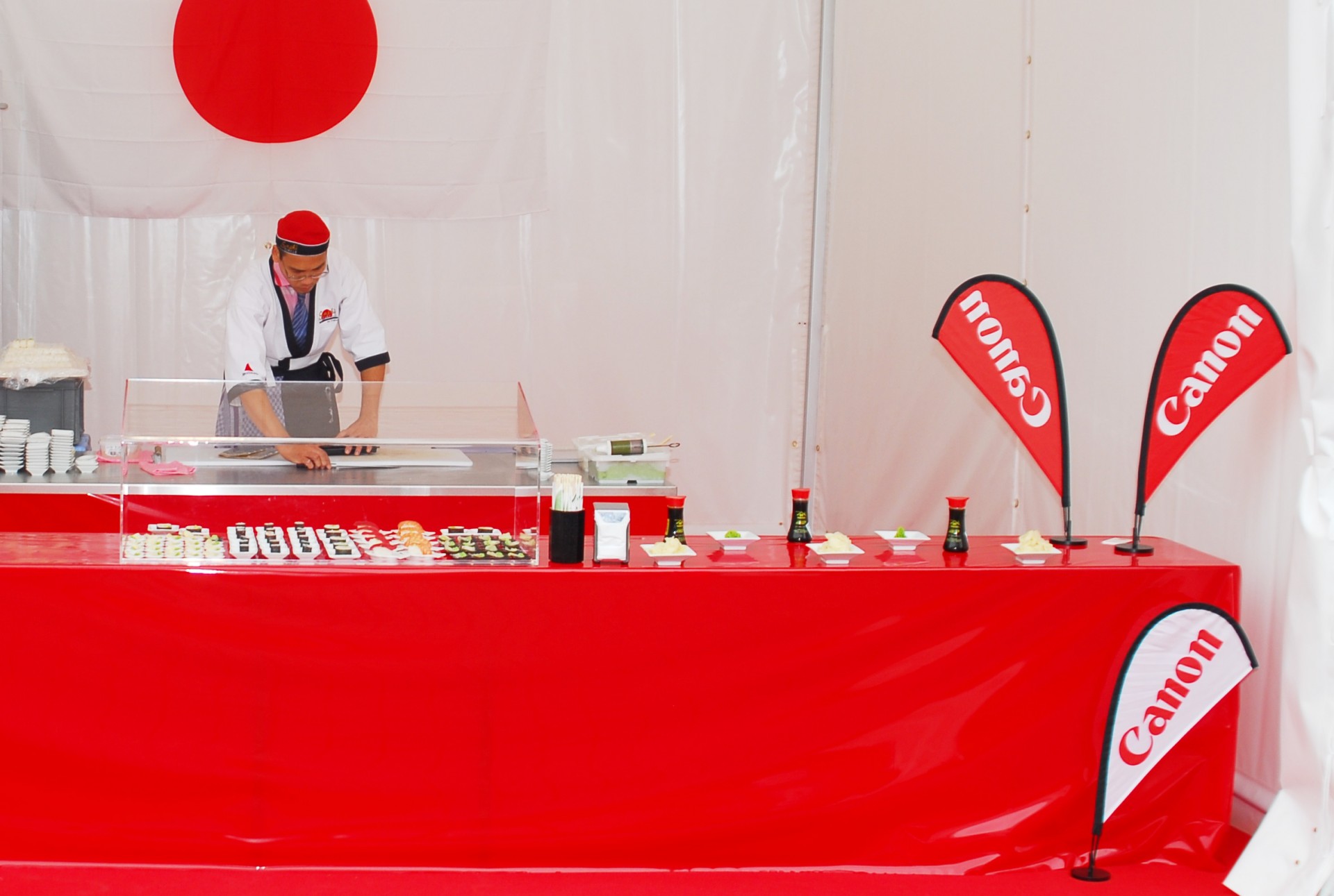 Ein Sushi-Stand mit rotem Tisch und Canon-Flaggen. Ein Koch bereitet Sushi vor.